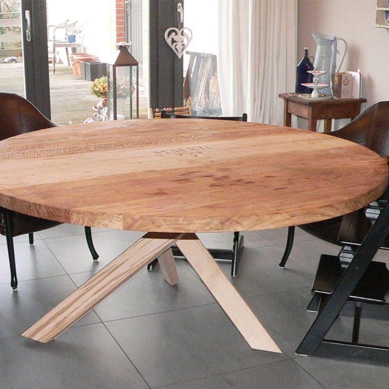 Maßgefertigter runder Esstisch aus Holz Ein großer, maßgefertigter runder Esstisch aus hellem Holz mit einzigartigem Design steht in einem modernen Esszimmer. Die Tischplatte ruht auf einem kreuzförmigen Holzgestell. Tisch nach Maß.