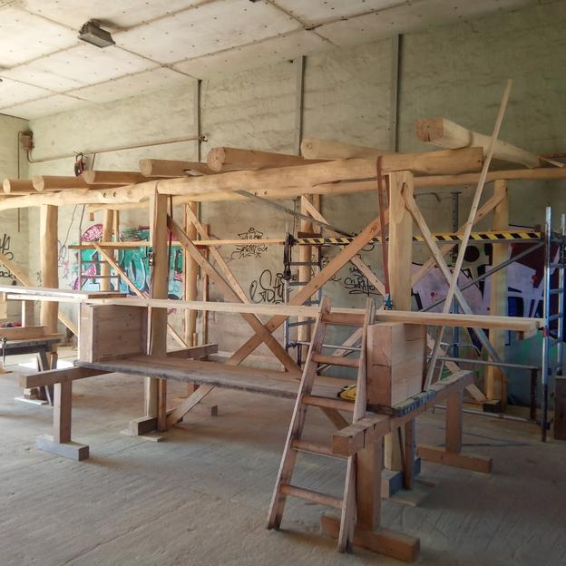 Unfertige Holzkonstruktion in einer Werkstatt, bestehend aus massiven Balken und Gerüsten, umgeben von Bauwerkzeugen und farbigen Graffiti-Wänden.
