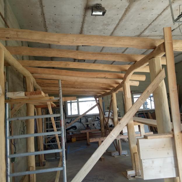 Aufbau eines Holzrahmenbaus in einer Werkstatt, mit massiven Balken und einer Leiter, umgeben von Graffiti und Bauutensilien.