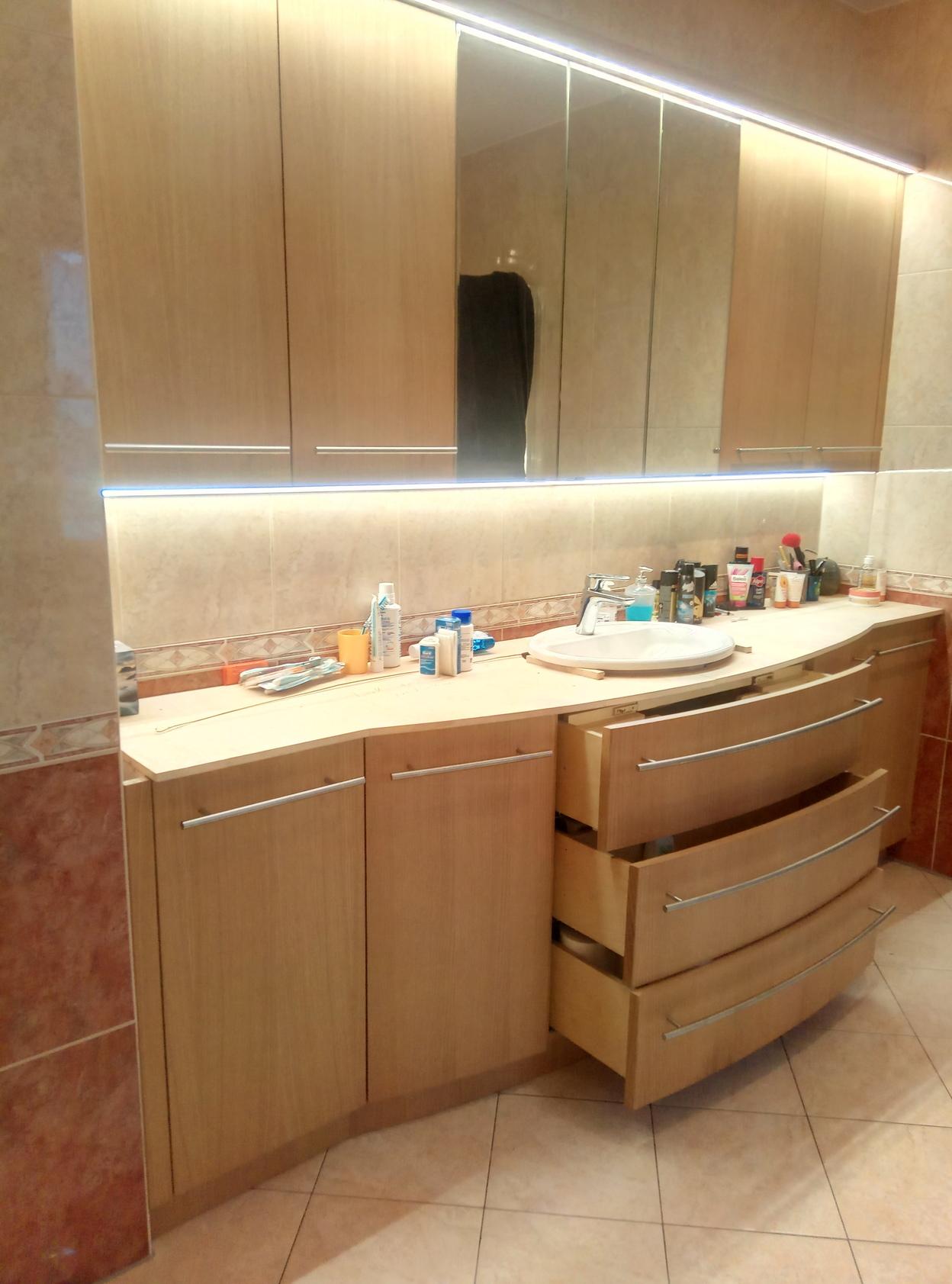 Moderne Badezimmerschranklösung mit geschwungenen Holzschubladen, LED-beleuchtetem Spiegel und viel Stauraum für Pflegeprodukte.