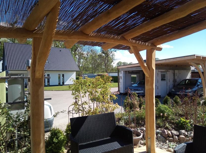 Gemütliche Terrasse mit einer Überdachung aus robusten Holzpfosten und Schattendach, umgeben von Pflanzen und mit Blick auf moderne Häuser und eine Garageneinfahrt.