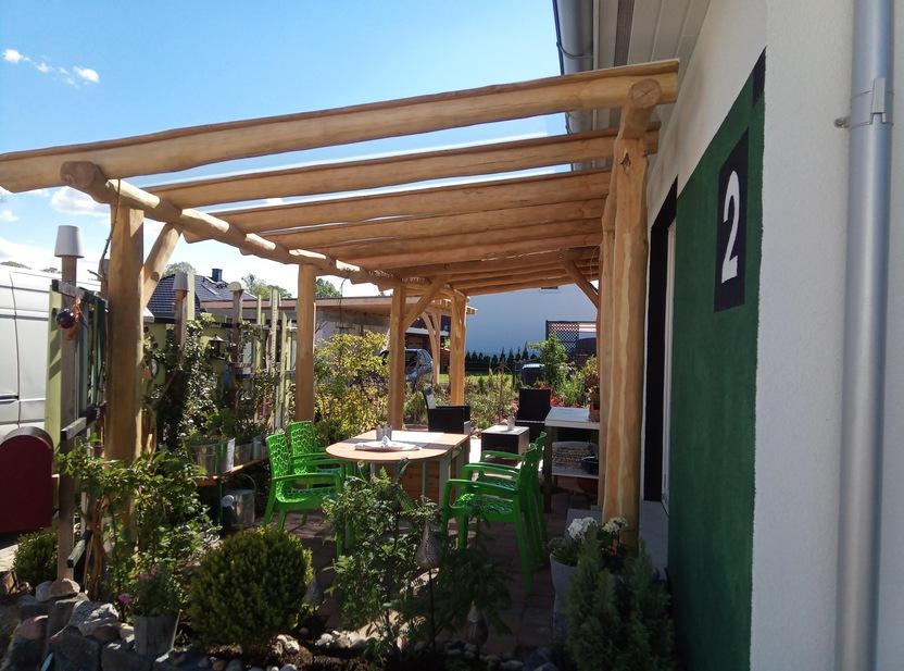 Terrasse mit natürlicher Holzüberdachung, grünen Gartenstühlen und einem Esstisch, umgeben von üppigen Pflanzen und einem gepflegten Gartenbereich.